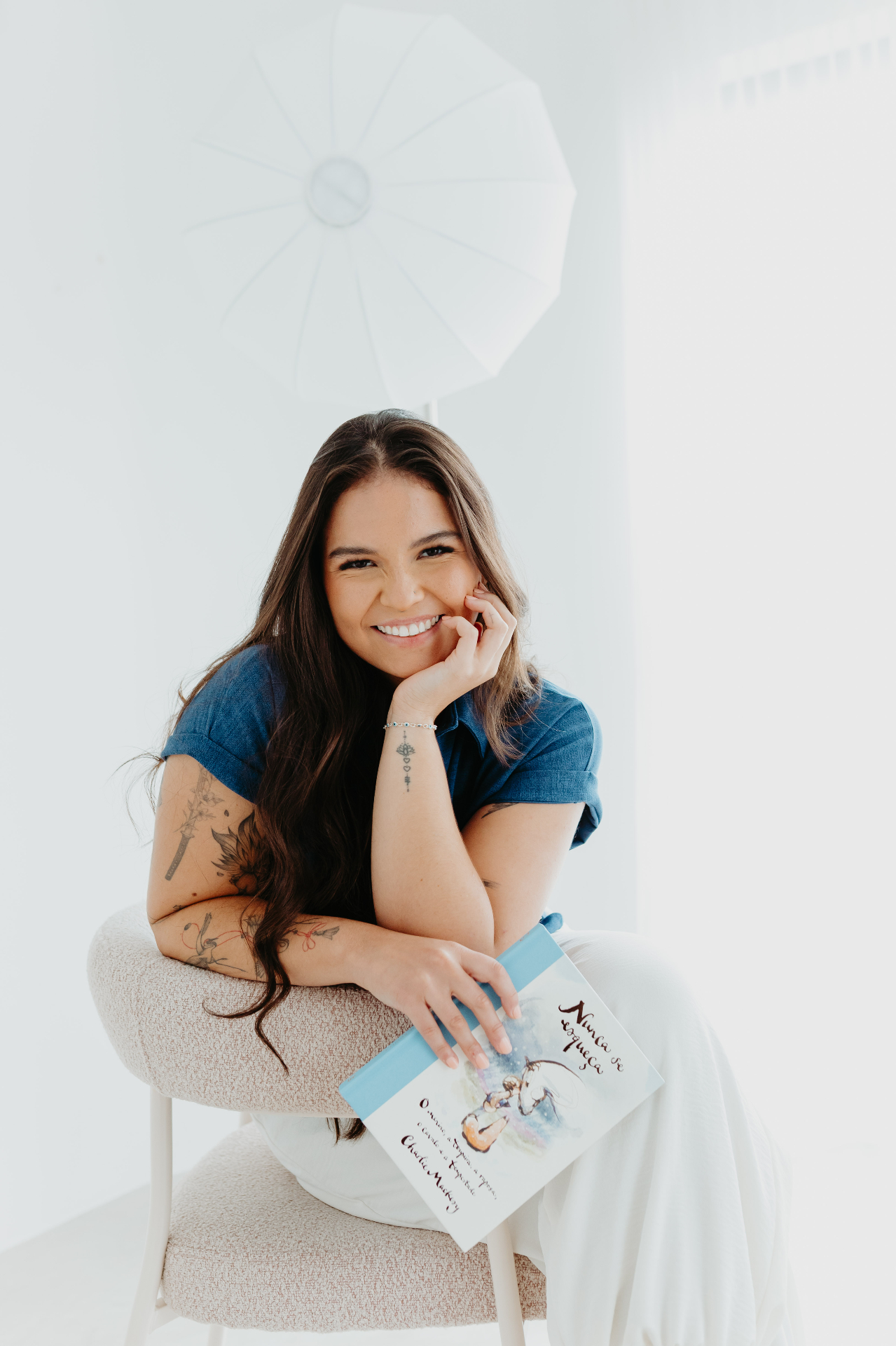Retrato de Kamila Tsuru sorrindo, apoiada em um assento claro, com um livro no colo.